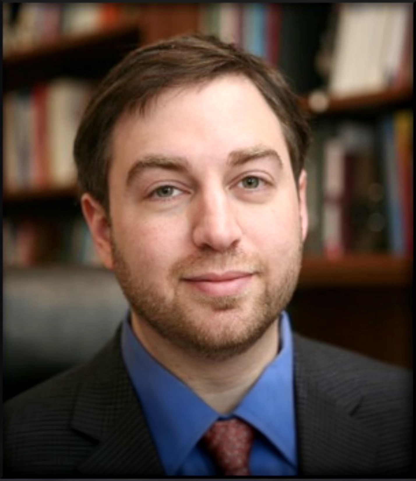 Dr. Aaron Goldner, in a jacket and tie, reading a book in front of a shelf of psychology texts